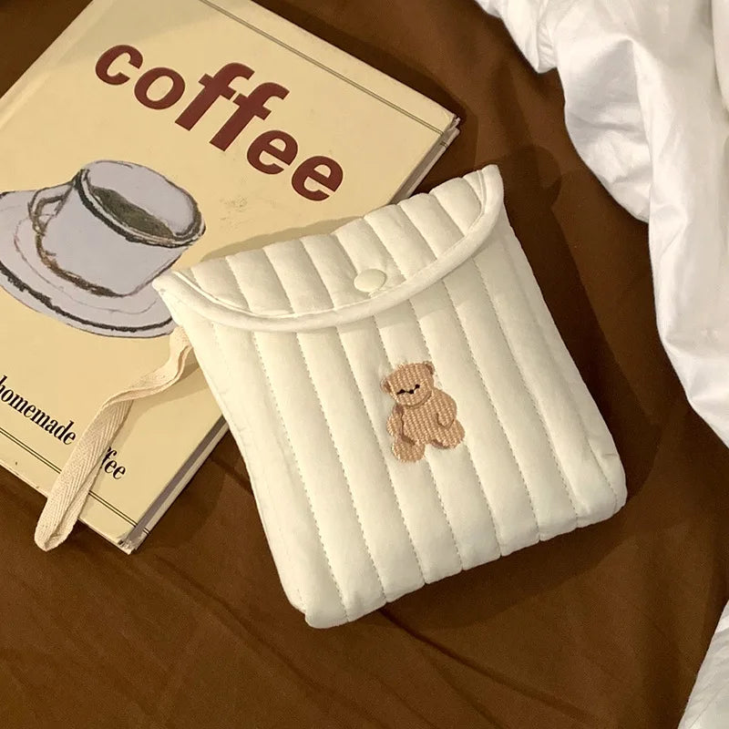 White quilted coffee cup cozy with a teddy bear patch on a brown surface with a coffee-themed book.